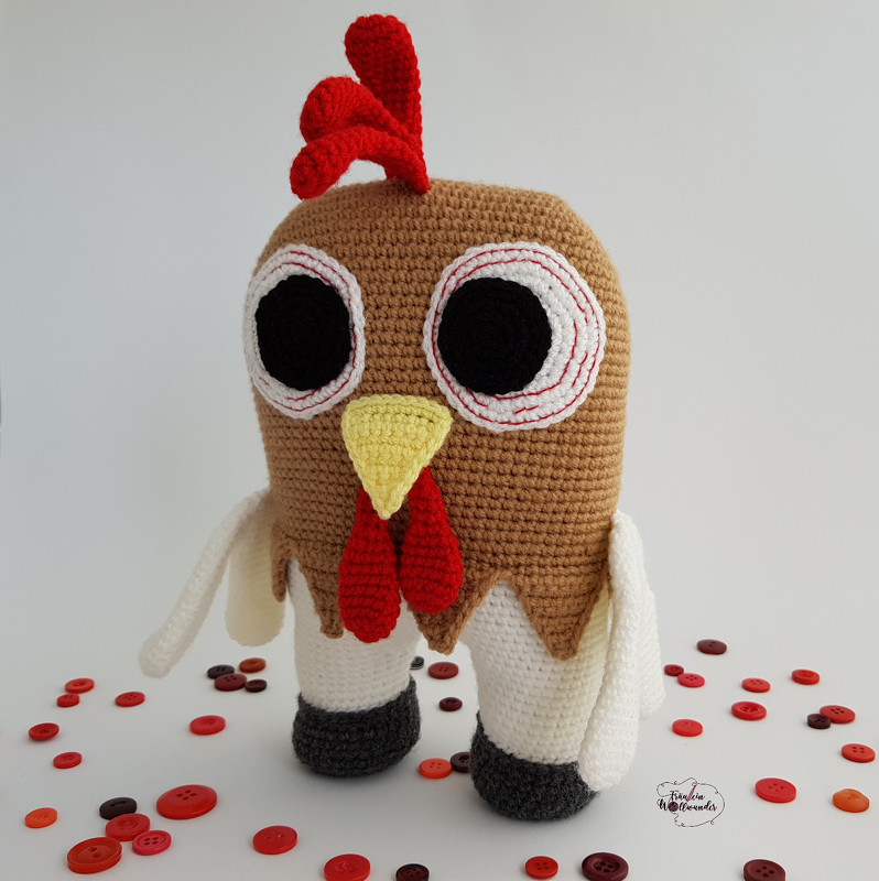 Gehäkeltes Amigurumi-Huhn mit braunem Körper, großen weißen Augen, gelbem Schnabel, rotem Kamm, weißen Beinen und dunklen Füßen