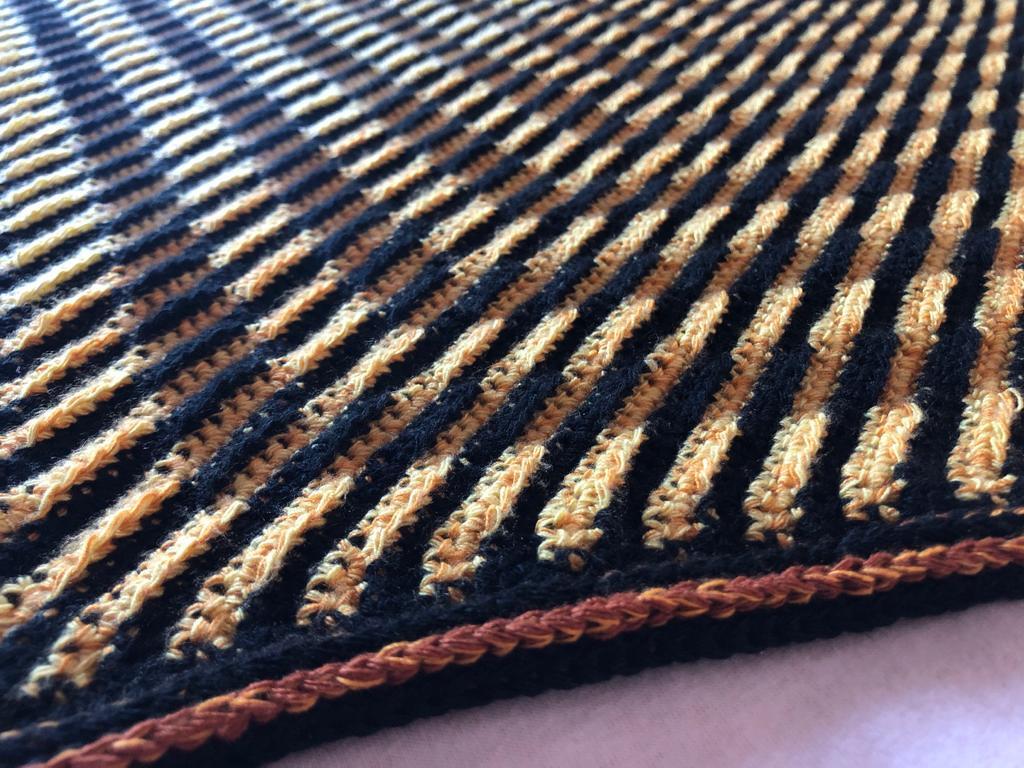 Nahaufnahme eines gehäkelten Dreieckstuchs mit schwarzen und ockergelben schmalen Streifen und rostfarbener Kante