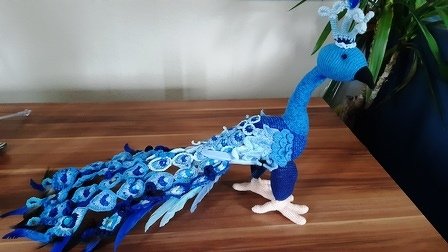Gehäkelter Pfau in Blautönen mit langem Schwanz und kleiner Krone auf Holzfläche