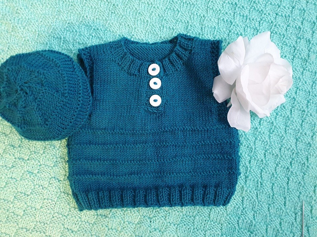 Petrolfarbenes gestricktes Baby-Oberteil mit passender Mütze und drei weißen Knöpfen auf mintgrüner Decke