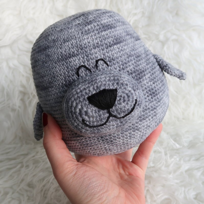 Hand hält einen gehäkelten grauen Robbenkopf mit schwarzer Nase und aufgesticktem Gesicht.