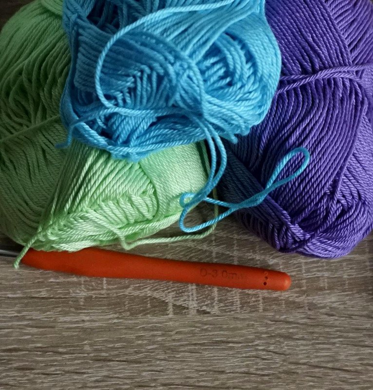 Mint green, aqua, and purple yarn skeins with an orange crochet hook on a wooden surface