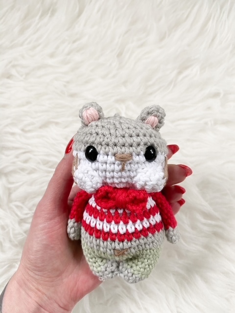 Hand hält kleines gehäkeltes Tier-Amigurumi mit rot-weißem Pullover vor weißem flauschigem Hintergrund