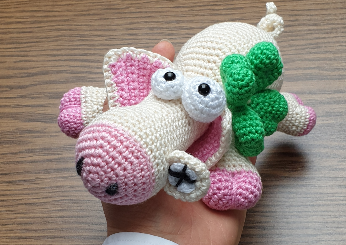 Gehäkeltes kleines Schwein in Creme und Rosa mit grünem Kleeblatt in einer Hand