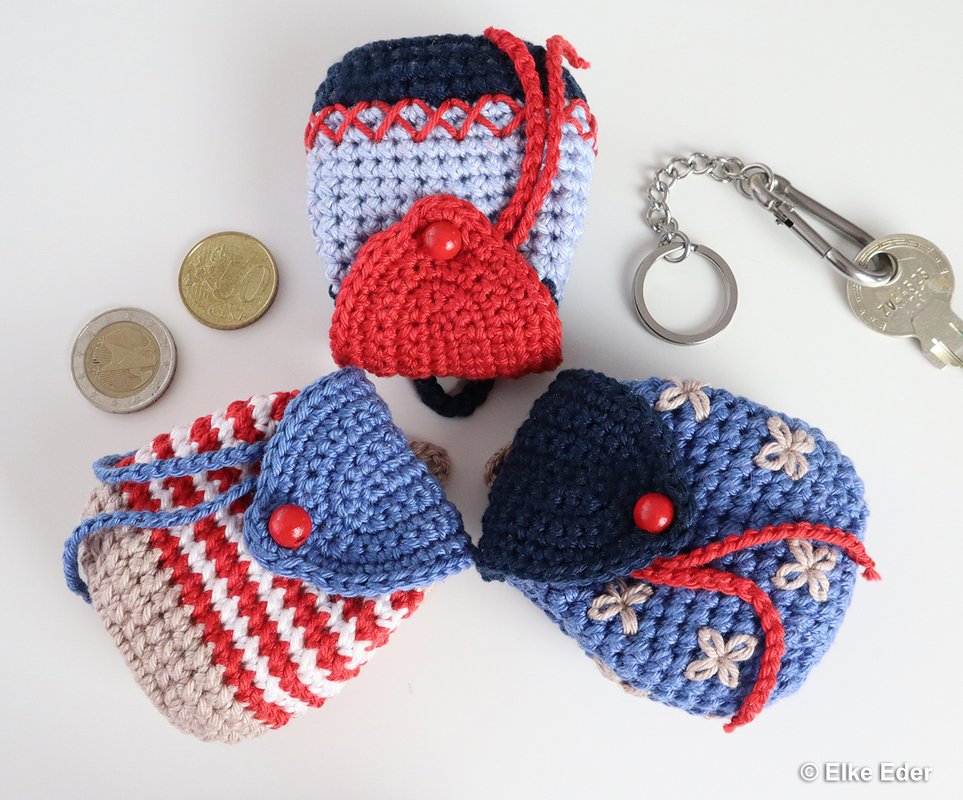 Three small crocheted coin pouch keychains in blue, red and striped colors with red button flaps, a metal keyring and coins on a white background