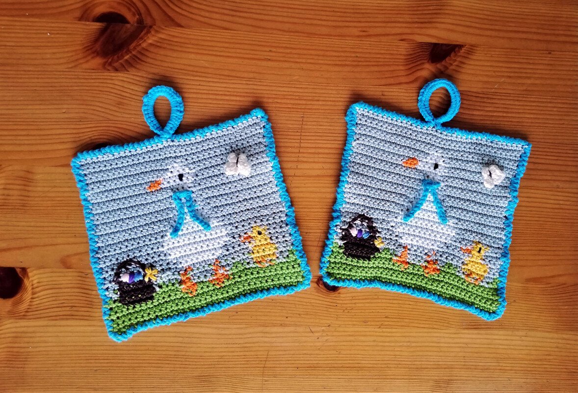 Zwei farbenfrohe gehäkelte Topflappen mit Vogel- und Kükenmotiven, hellblauer Umrandung und Aufhängeschlaufen