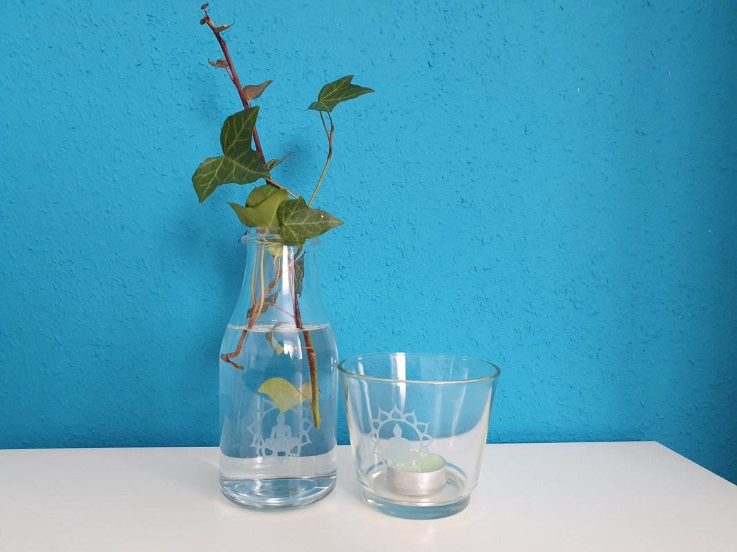 Zwei Glasgefäße (Flasche mit Efeu, Teelichtglas) verziert mit weißem Meditationsmotiv vor blauem Hintergrund