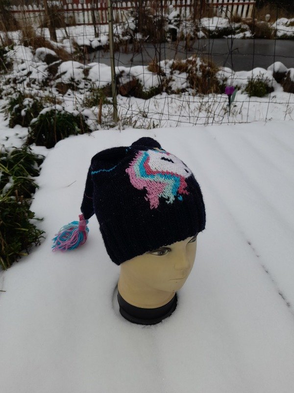 Dunkelblaue gestrickte Kindermütze mit pastellfarbenem Einhornmotiv und Bommel auf Schaufensterkopf im Schnee