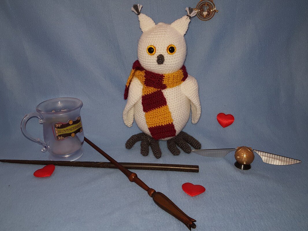 Weißes gehäkeltes Eulen‑Amigurumi mit gelb‑rotem gestreiftem Schal, umgeben von Zauberstäben, einer goldfarbenen Kugel mit Flügeln, einer transparenten Tasse und roten Herzen auf blauem Hintergrund.
