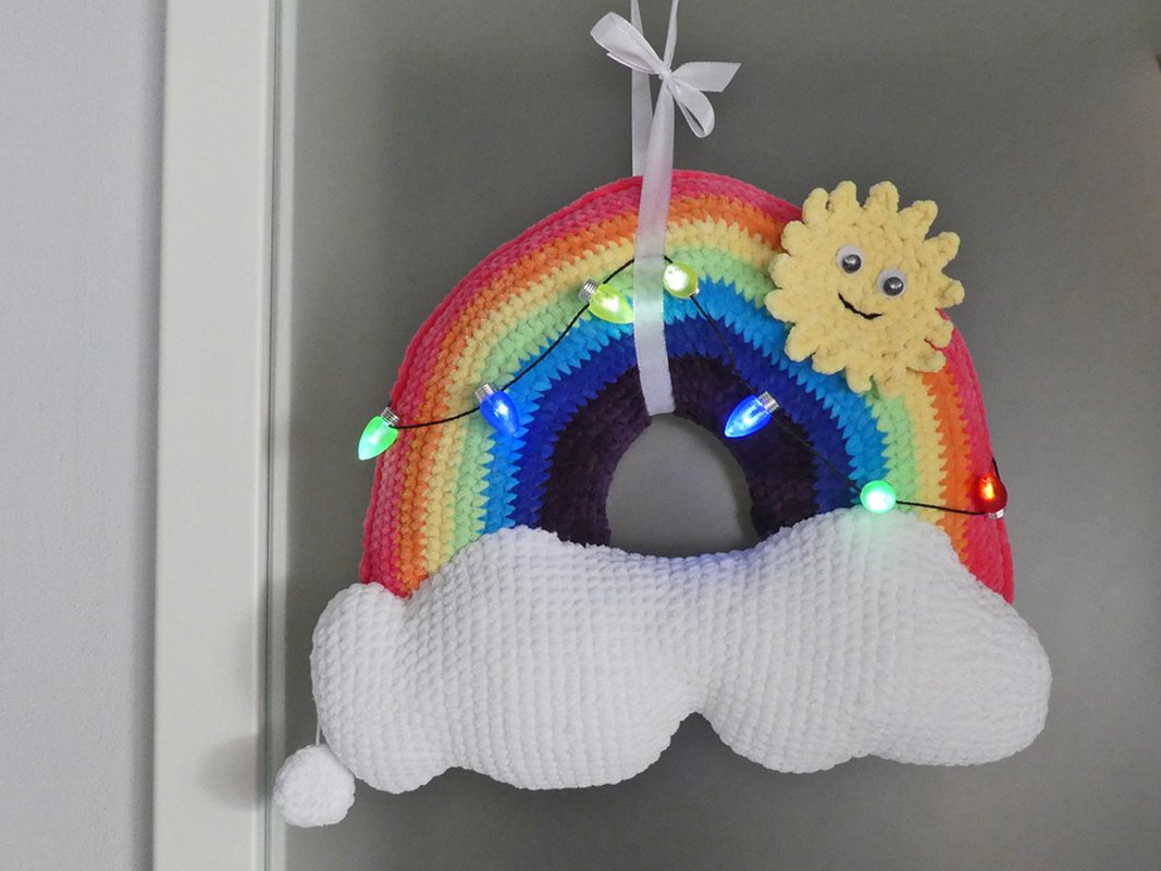 Gehäkelter Regenbogen mit weißer Wolke, gelber Sonne mit Wackelaugen, umwickelt von bunten Lichterketten, an weißem Band hängend.