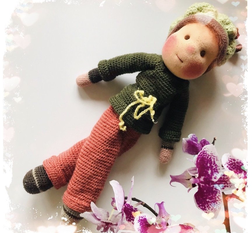 Gehäkelte Amigurumi-Puppe liegend mit grünem Pullover, rostroter Hose und Mütze neben Orchideen