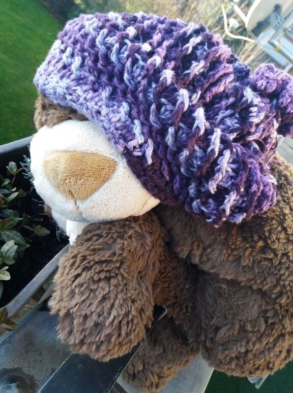Lila mehrfarbige gehäkelte Slouch‑Mütze auf braunem Kuscheltier