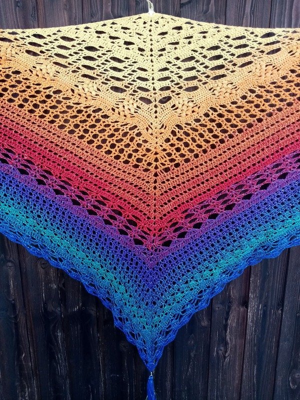 Gehäkeltes Dreieckstuch mit mehrfarbigem Farbverlauf und Lochmuster vor dunklem Holz hängend.