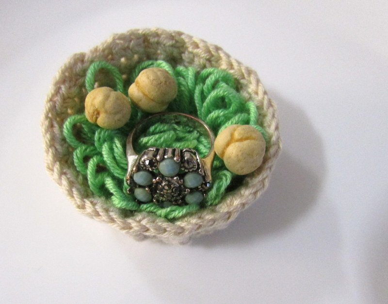 Handgehäkeltes Nest in Beige mit grünem Garn, drei kleinen Dekoeiern und einem Ring mit hellblauen Akzenten.