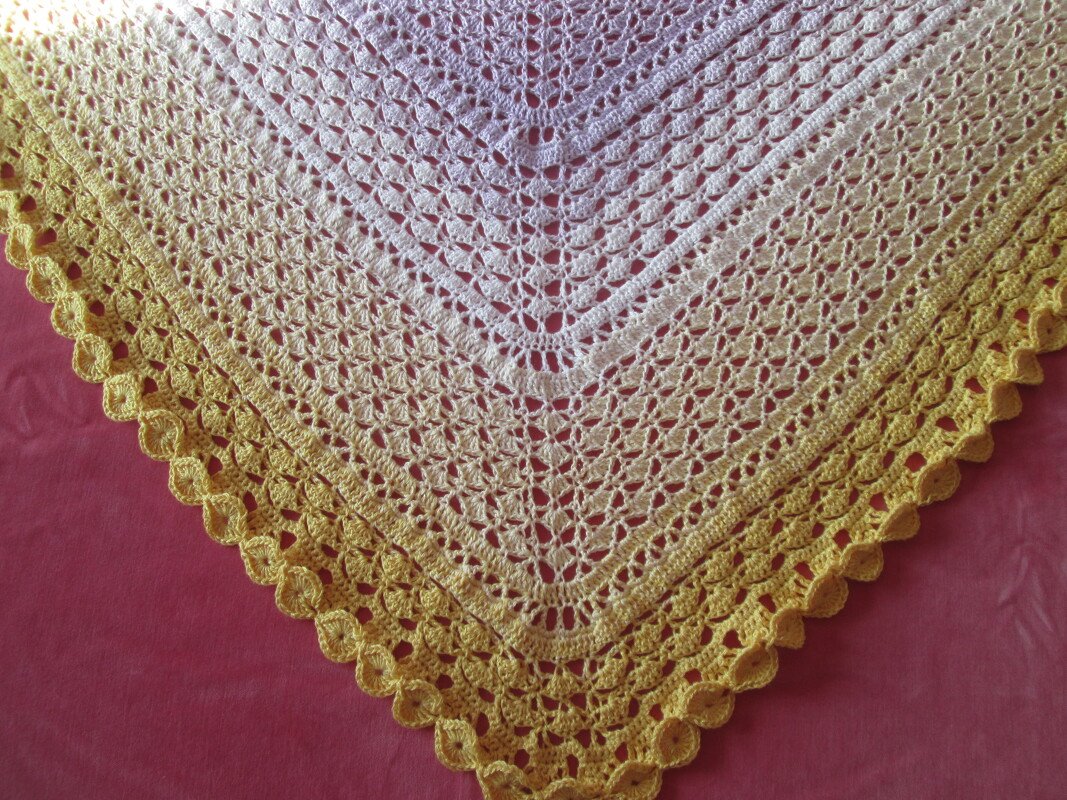 Detail eines gehäkelten Dreieckstuchs in Creme- bis Gelbtönen mit Lochstäbchen, Fächermotiven und breiter Muschelkante auf rotem Untergrund