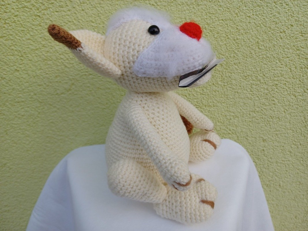 Beiges gehäkeltes Amigurumi-Haustier mit roter Nase, seitlich sitzend vor hellgrüner Wand