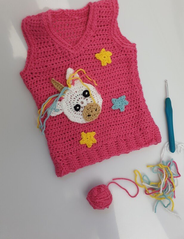 Pink crocheted children's sleeveless vest with unicorn face and star appliques beside a crochet hook and yarn.