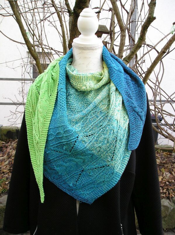 Gestricktes Dreieckstuch in Grün- und Blautönen mit Zopf- und Lochmuster auf einer Schneiderpuppe.