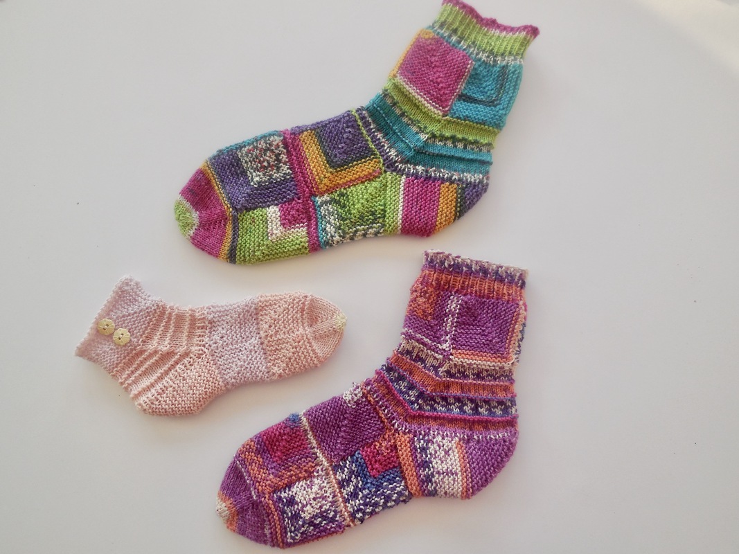 Drei gestrickte Kindersocken: eine rosa Babysocke mit Knöpfen und zwei bunte Mustersocken auf weißem Grund.