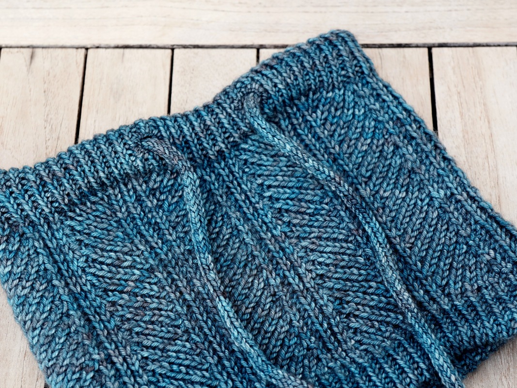 Gestrickter Loopschal in Blau-Grau mit Fischgrätmuster und Kordelzug auf hellem Holz