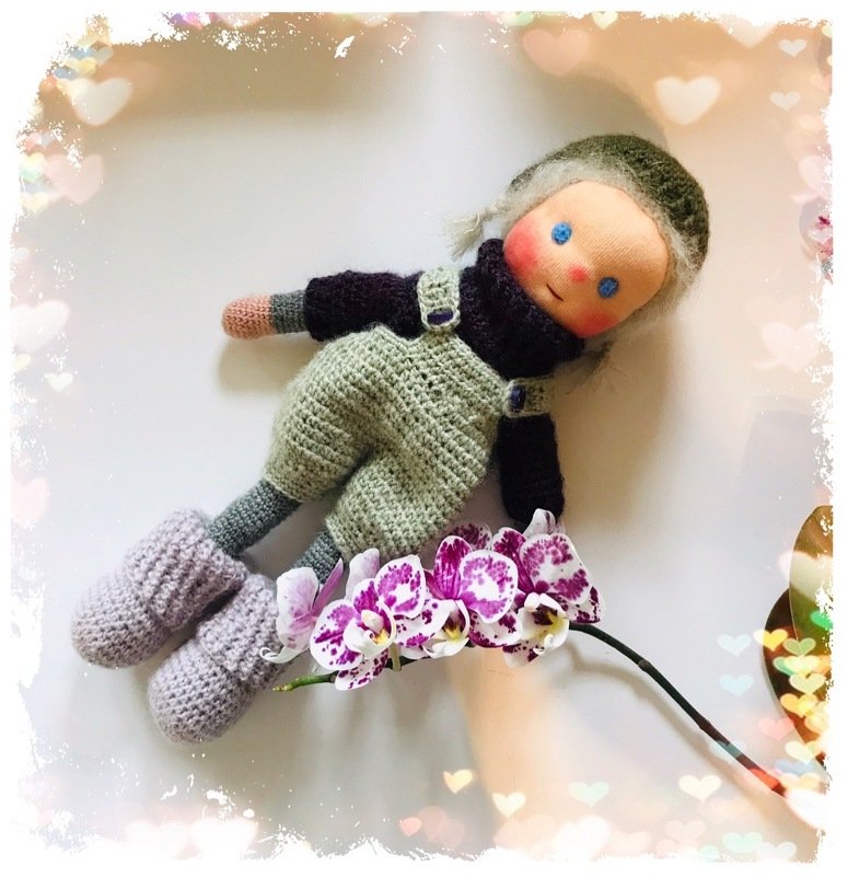Gehäkelte Amigurumi-Puppe mit Mütze, Latzhose und Stiefeln neben Orchideen