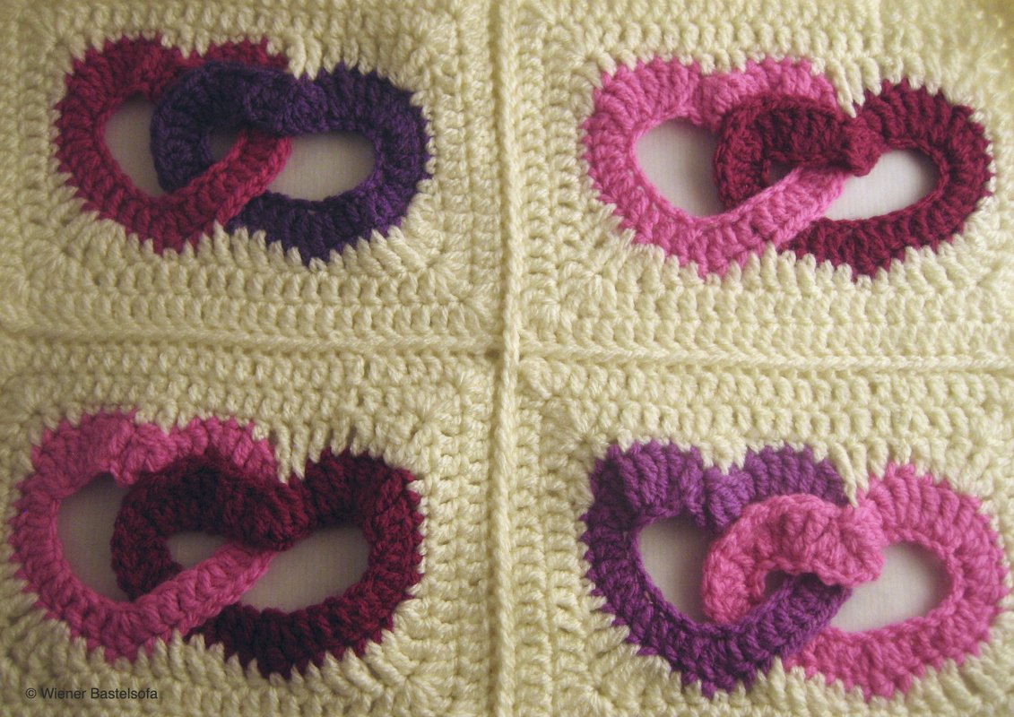 Vier gehäkelte Quadrate mit cremefarbenem Hintergrund und ineinander verschlungenen Herz-Applikationen in Pink und Violett