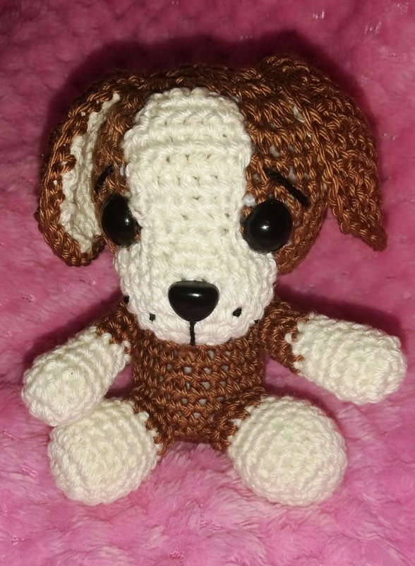 Handgehäkelter sitzender Amigurumi‑Hund in Braun und Weiß mit schwarzen Augen auf pinkfarbenem, weichem Untergrund