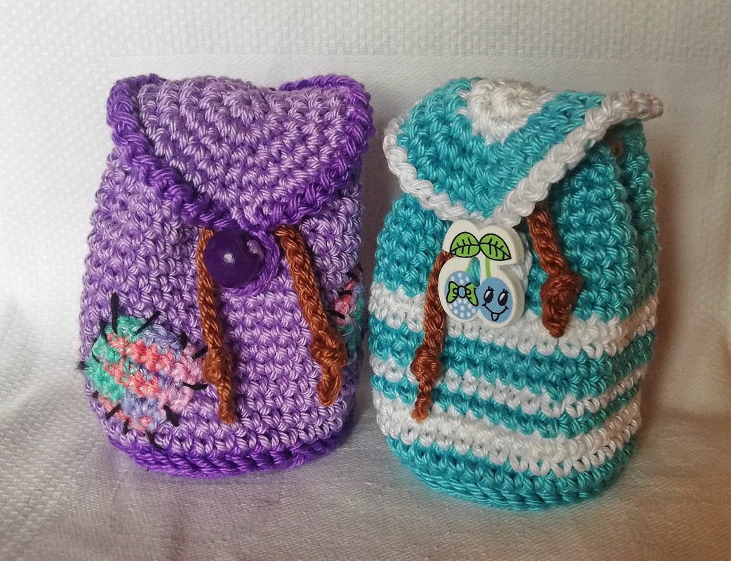 Nahaufnahme von zwei gehäkelten Mini-Rucksäcken: links lila mit mehrfarbiger Verzierung, rechts türkis-weiß gestreift mit Kirsch-Knopf mit Gesicht.