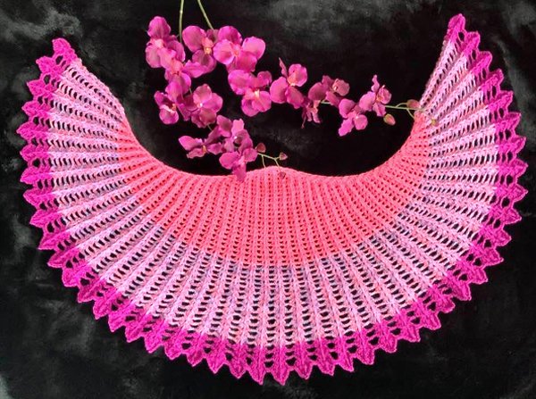 Gehäkeltes halbrundes Tuch in Pink‑Farbverlauf mit Zackenrand auf schwarzem Hintergrund, daneben rosa Orchideen