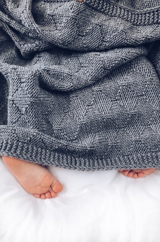 Gray knitted blanket with geometric textured stitch and ribbed edge draped over white bedding with bare feet peeking out