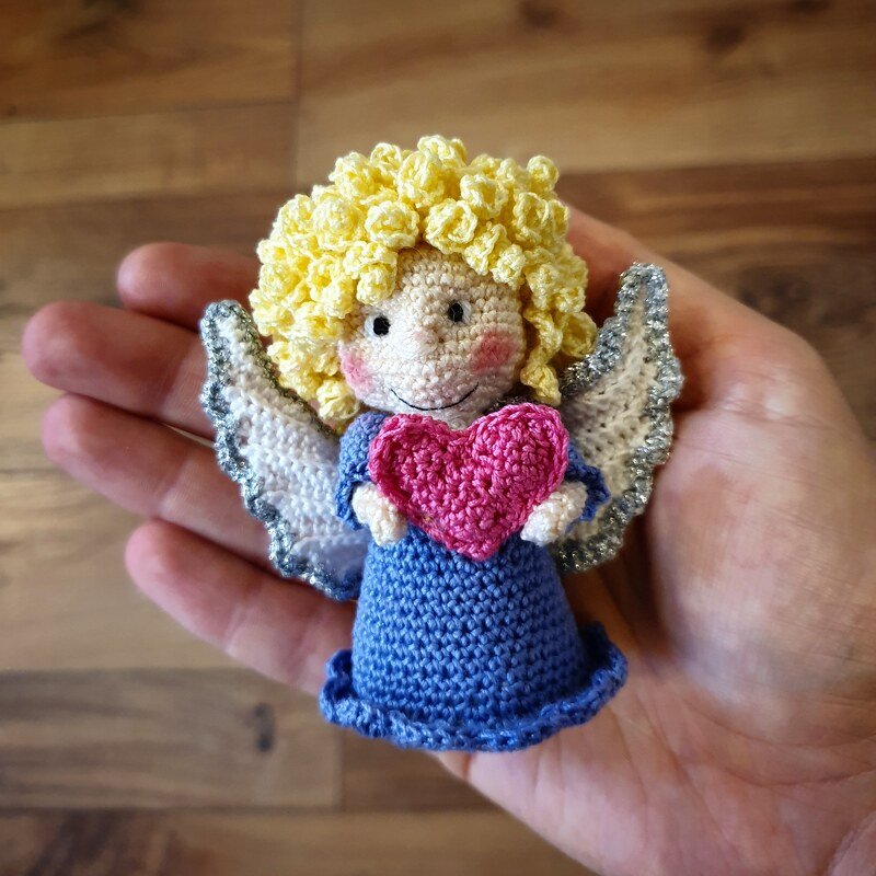 Amigurumi-Häkelengel mit gelbem Lockenkopf, blauem Kleid und rosa Herz, gehalten in einer Hand.