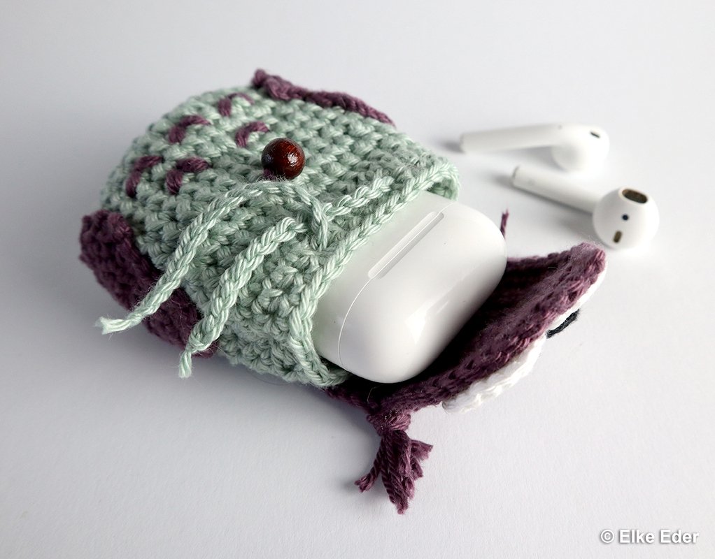 Mint and purple crocheted owl pouch with a white earbud charging case partly inside and two white earbuds nearby.