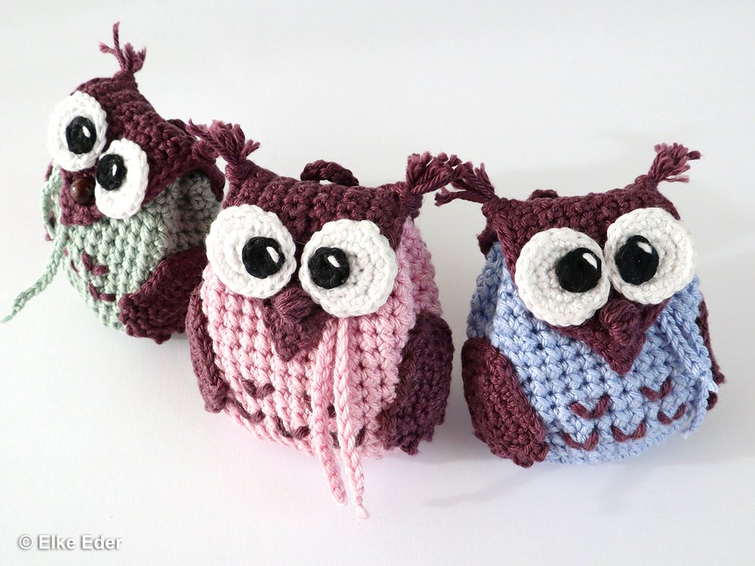 Three small crocheted owl keychains in pastel green, pink, and blue with large white-and-black eyes on a white surface.