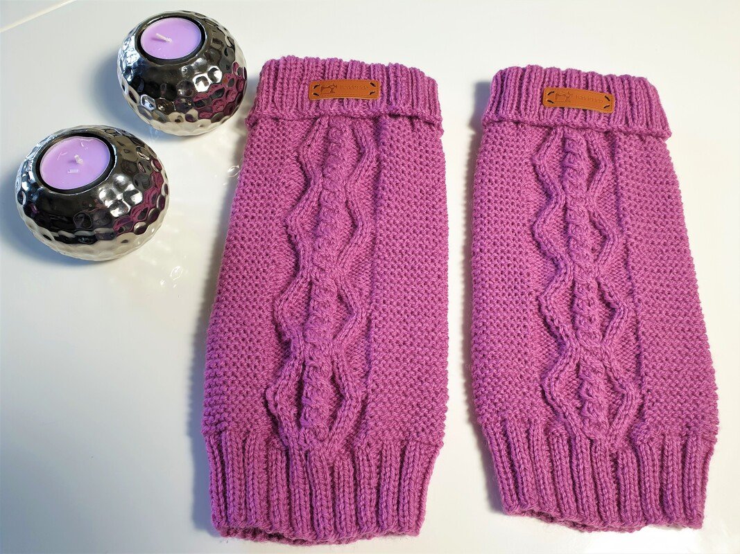 Zwei lila gestrickte Beinstulpen mit Zopfmuster auf weißem Untergrund neben zwei silbernen Kerzenhaltern mit lila Teelichtern.