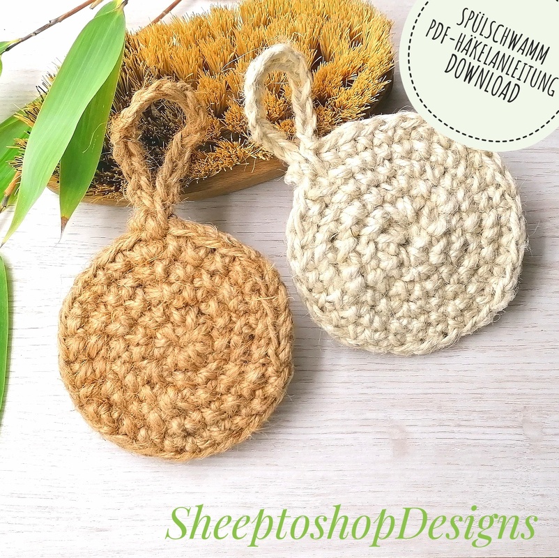 Zwei runde, gehäkelte Spülschwämme in Naturtönen mit Aufhängeschlaufen auf hellem Holztisch neben einer Holzbürste und grünen Blättern.