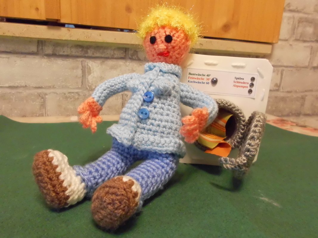 Gehäkelte Puppe mit gelbem Haar, hellblauem Jäckchen und braunen Schuhen neben einer Spielzeug-Waschmaschine