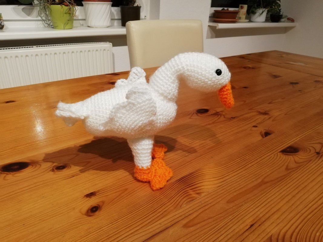 Gehäkelte weiße Gans mit orangefarbenem Schnabel und Füßen auf braunem Holztisch.