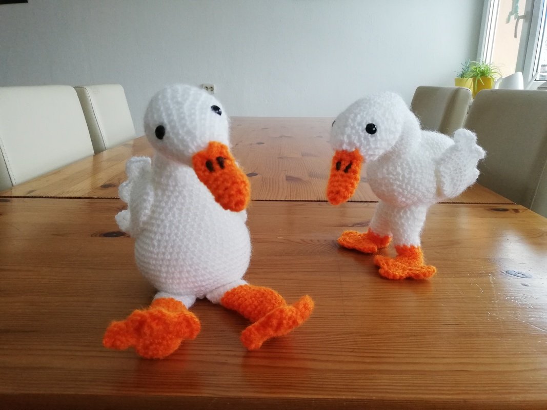 Zwei weiße gehäkelte Enten-Amigurumi mit orange Schnäbeln und Füßen auf Holztisch