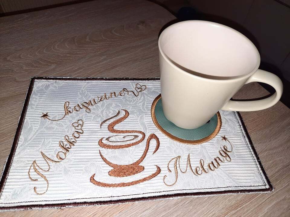 Rechteckiger bestickter Untersetzer mit Kaffeetassen-Motiv und den Schriftzügen 'Mokka', 'Kapuziner', 'Melange', darauf eine weiße Tasse