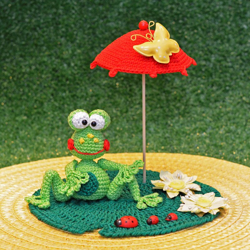Gehäkelter grüner Frosch auf Seerosenblatt mit rotem Sonnenschirm und Seerosen