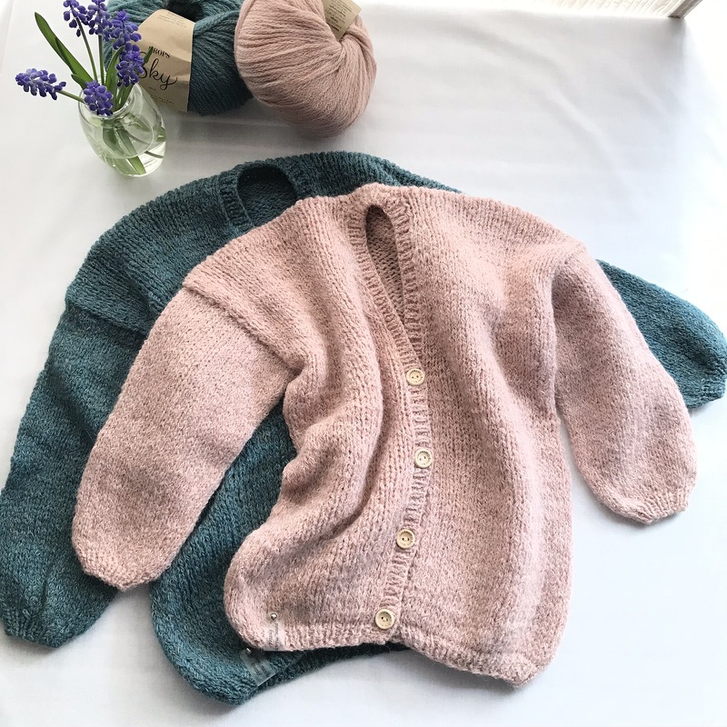 Zwei gestrickte Kinderjacken, rosa Jacke mit Knöpfen liegt auf türkiser Jacke, Garnknäuel und kleine Vase daneben