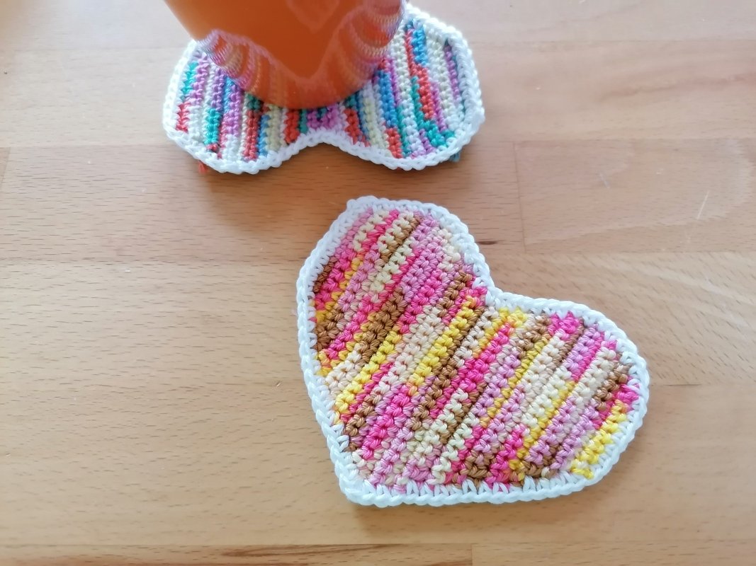 Zwei bunte herzförmige gehäkelte Untersetzer auf Holz, einer unter einer orangefarbenen Tasse