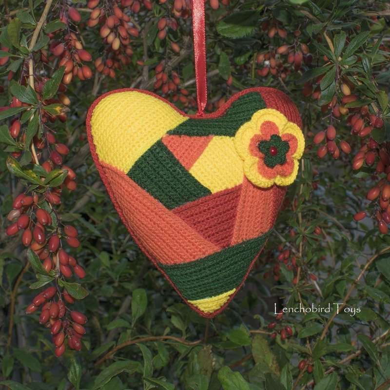 Crocheted patchwork heart ornament in yellow, orange and green with a small flower, hanging by a ribbon against leafy branches