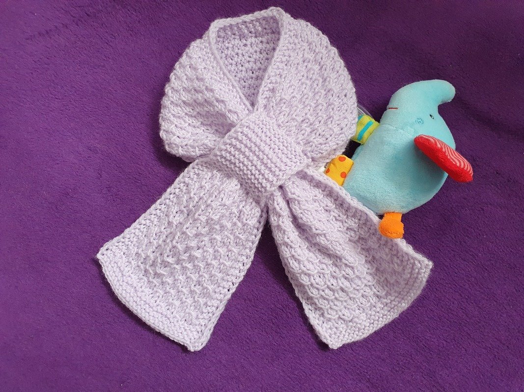 Gestrickter Babyschal in hellem Lila mit Schlitz zum Durchziehen, auf violettem Hintergrund neben einem bunten Stofftier.