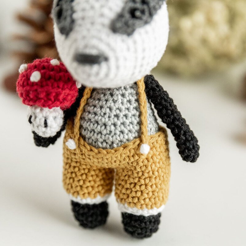 Gehäkeltes Amigurumi‑Tier in senffarbener Latzhose hält roten Pilz