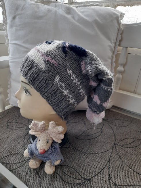 Gestrickte Kindermütze in Grau, Weiß, Rosa und Dunkelblau mit langem Zipfel und rosa Bommel auf einer Schaufensterpuppe.