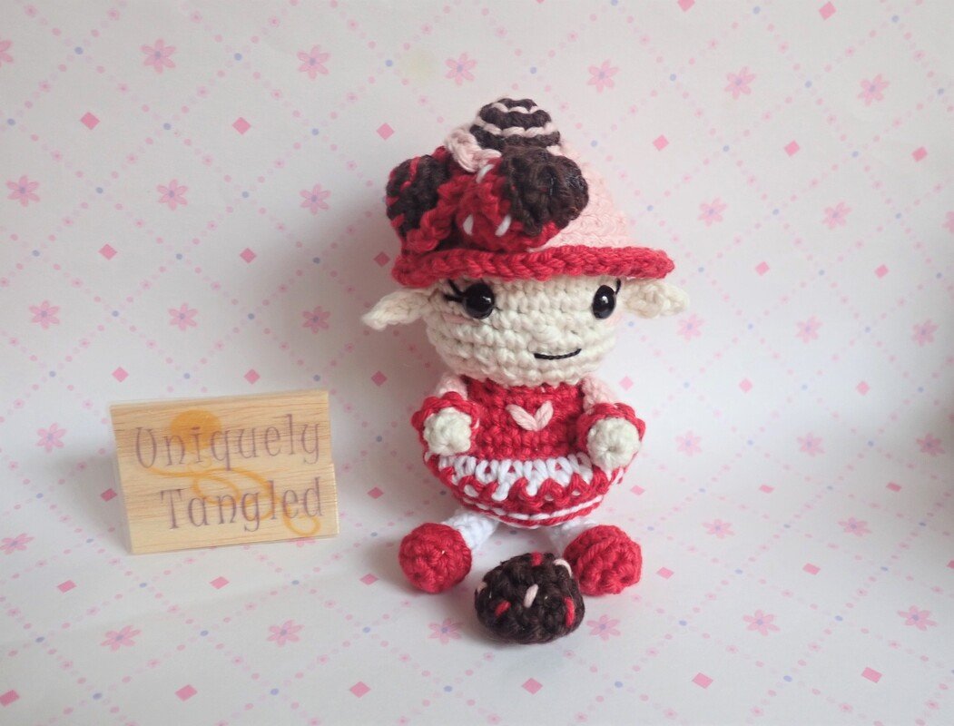 Red-and-white amigurumi lamb wearing a dress and matching hat with a small crocheted ball in front.