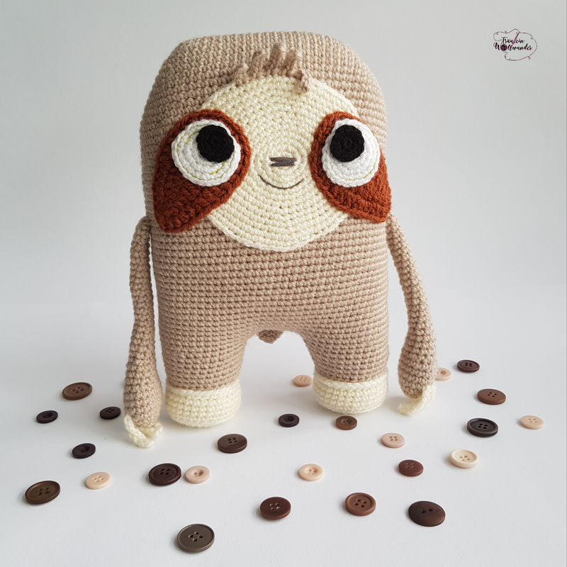 Gehäkeltes beige-braunes Amigurumi-Faultier mit großen Augen vor hellem Hintergrund, umgeben von verstreuten Knöpfen
