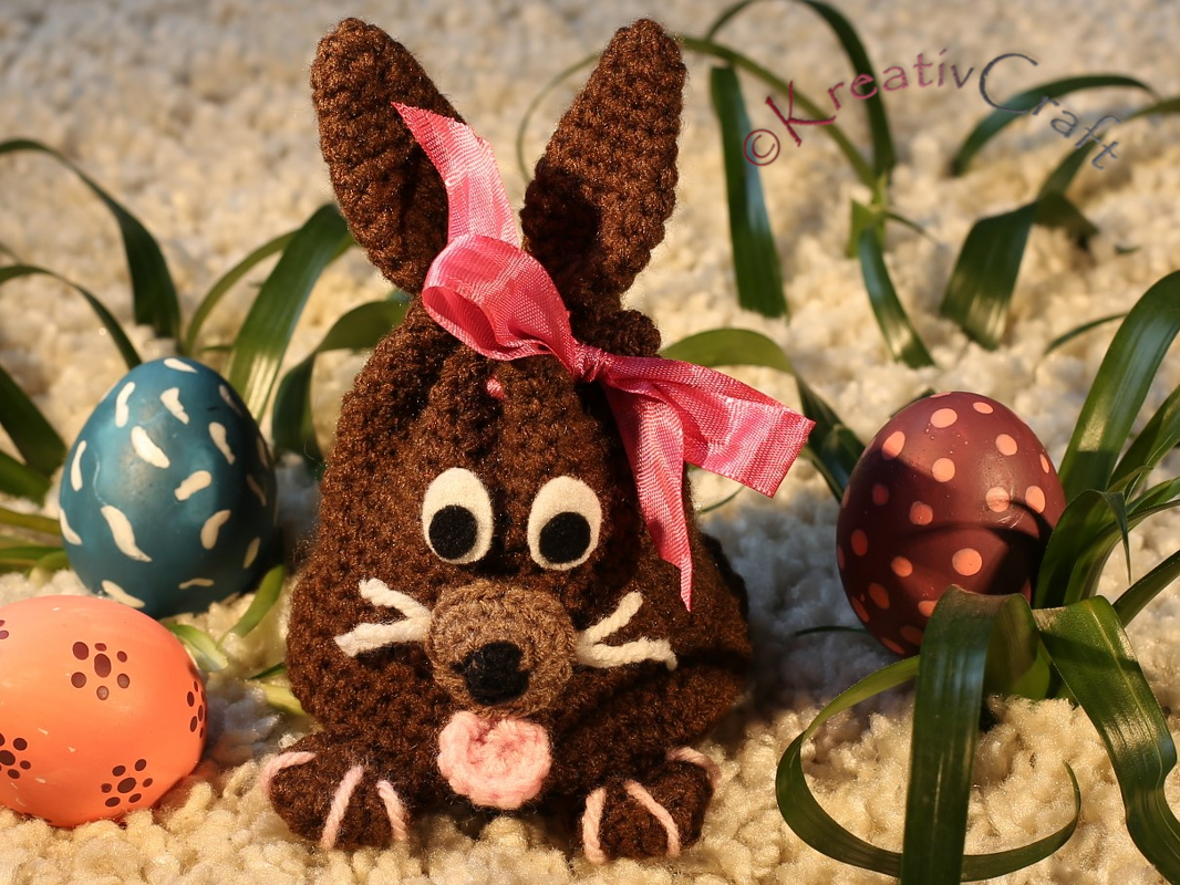 Brauner gehäkelter Amigurumi-Hase mit rosa Schleife, umgeben von drei bemalten Ostereiern und grünen Blättern auf weißem, flauschigem Untergrund.