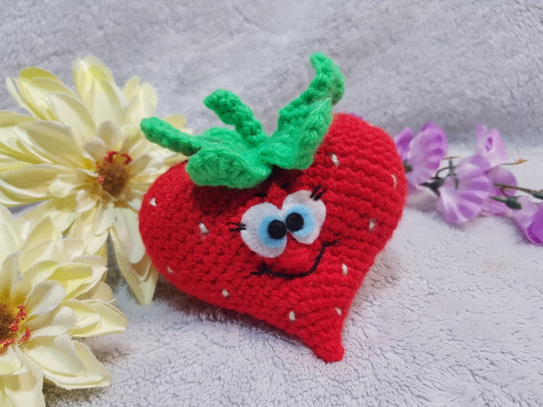 Gehäkeltes rotes Erdbeer‑Herz Amigurumi mit grünen Blättern, weißen Samenstichen und lächelndem Gesicht auf hellem Stoff